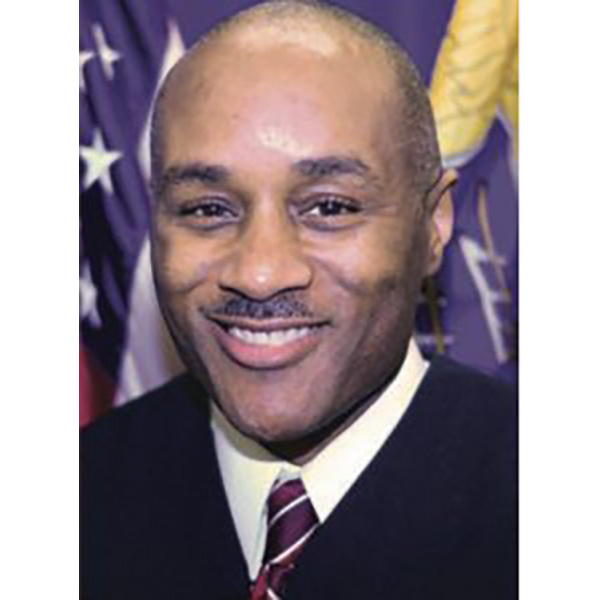 Rodney thompson in a black suit and tie smiling at the camera in front of the american flag